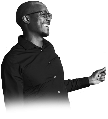 Smiling man wearing glasses in a black button-down shirt, facing left with an outstretched arm as if gesturing or inviting.